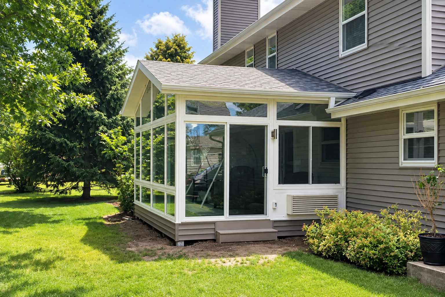 Custom sunroom with white-framed glass walls and a gabled roof extending from a gray-sided house.