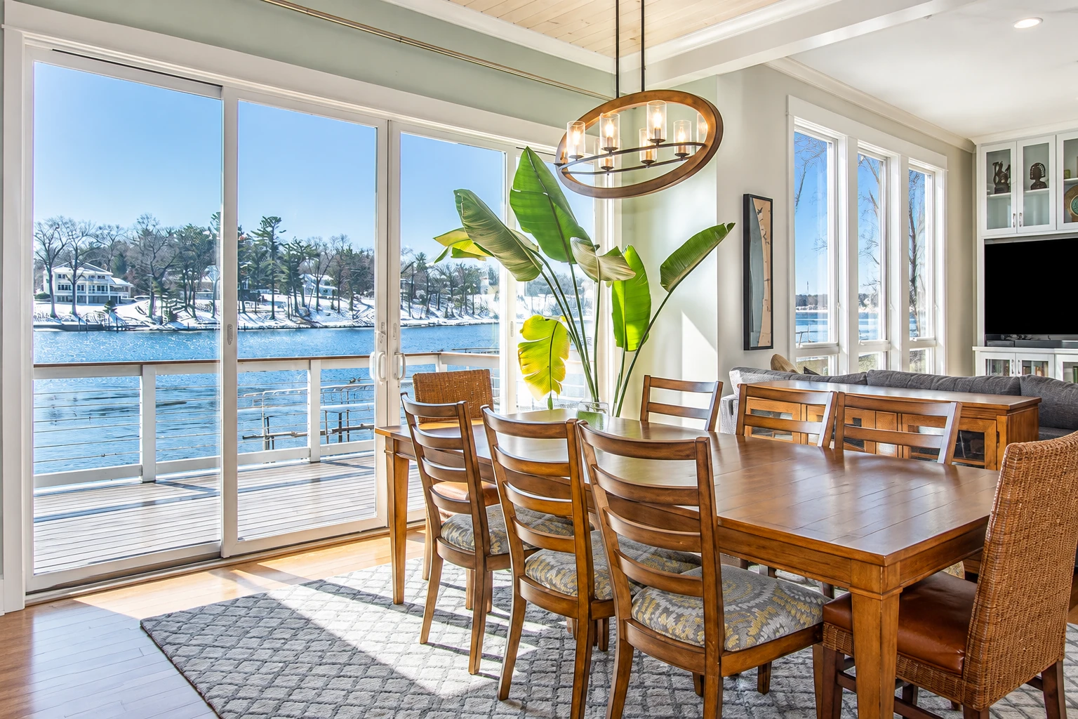 A bright dining room with large windows and sliding glass doors overlooking a snowy waterfront, furnished with a wooden table and chairs.