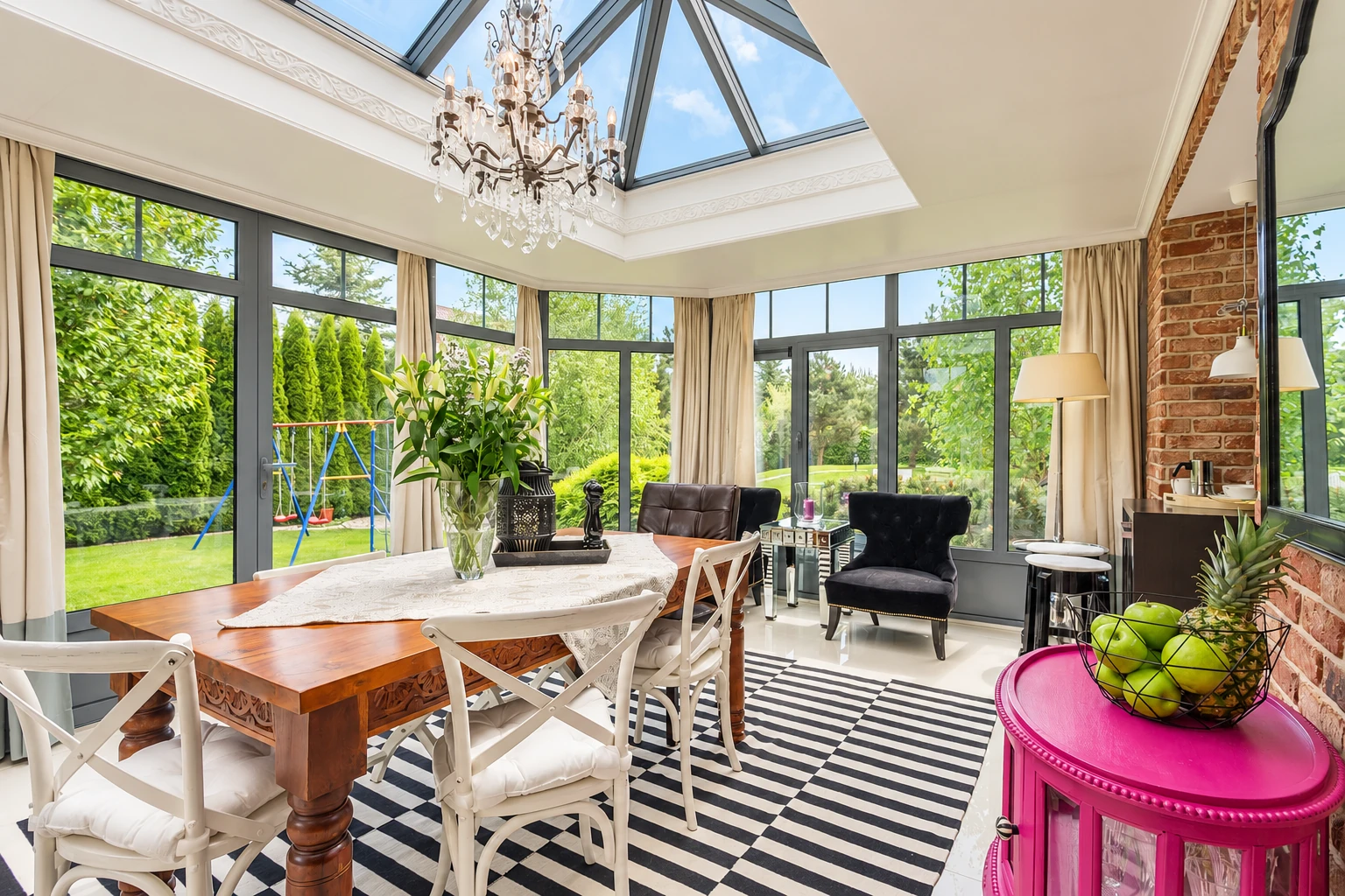 Four-season sunroom with a glass roof, chandelier, dining table, and large windows facing a garden.
