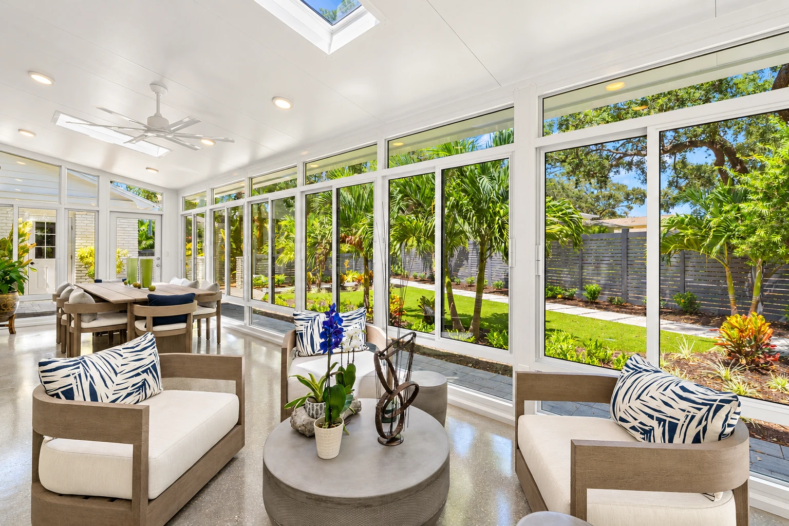 Four-season sunroom with white-framed windows, skylights, and lounge seating overlooking a sunny tropical garden.