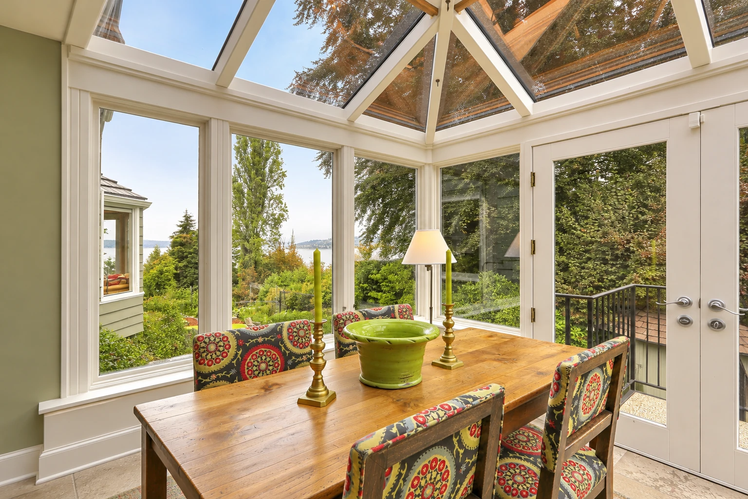 A glass-roofed solarium with white framing contains a wooden dining table and overlooks a leafy garden through tall windows.