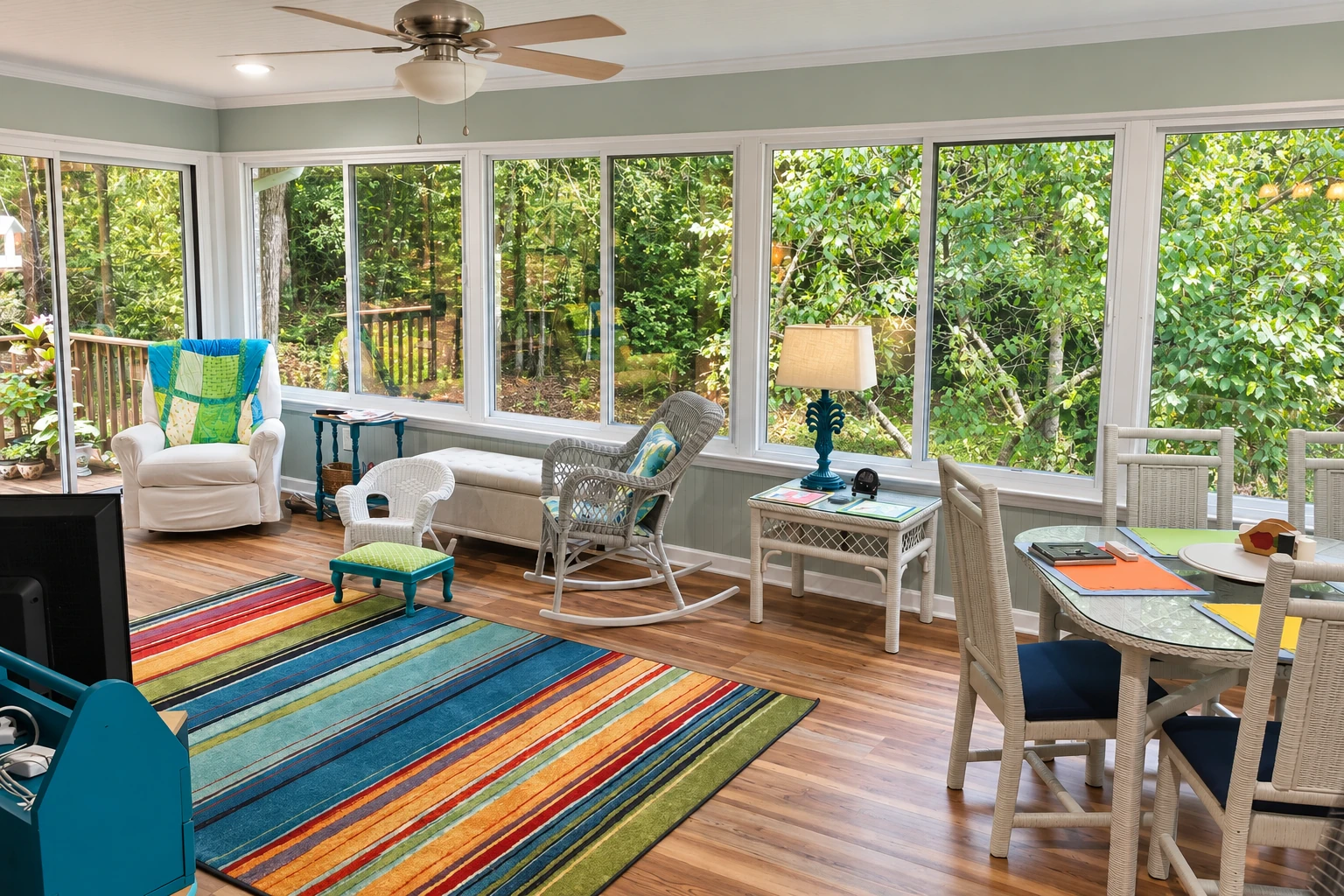 A bright sunroom with white-framed windows overlooking trees, furnished with wicker chairs, a dining table, and a colorful striped rug.
