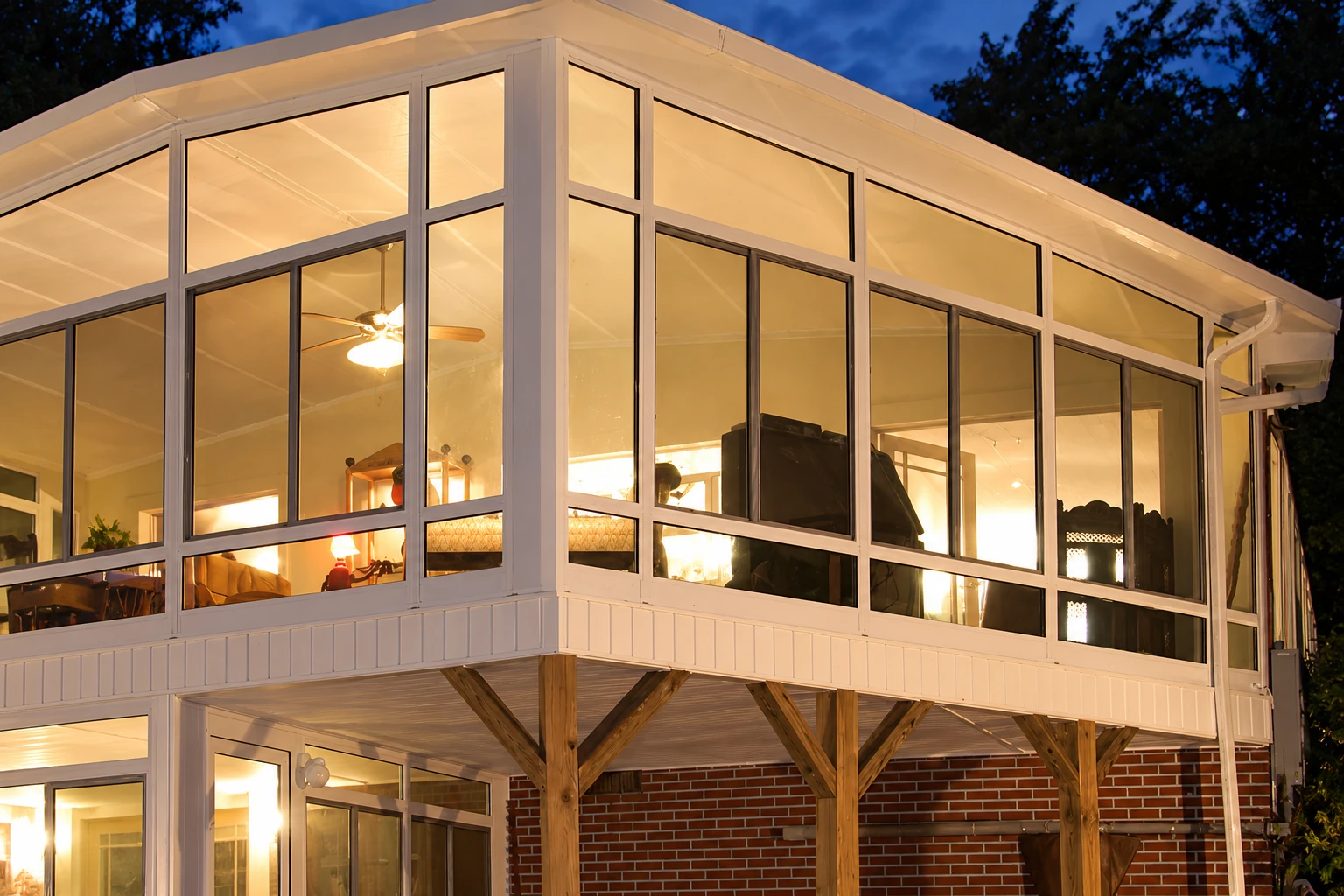 Illuminated three-season sunroom with tall white-framed windows on a raised deck at dusk.