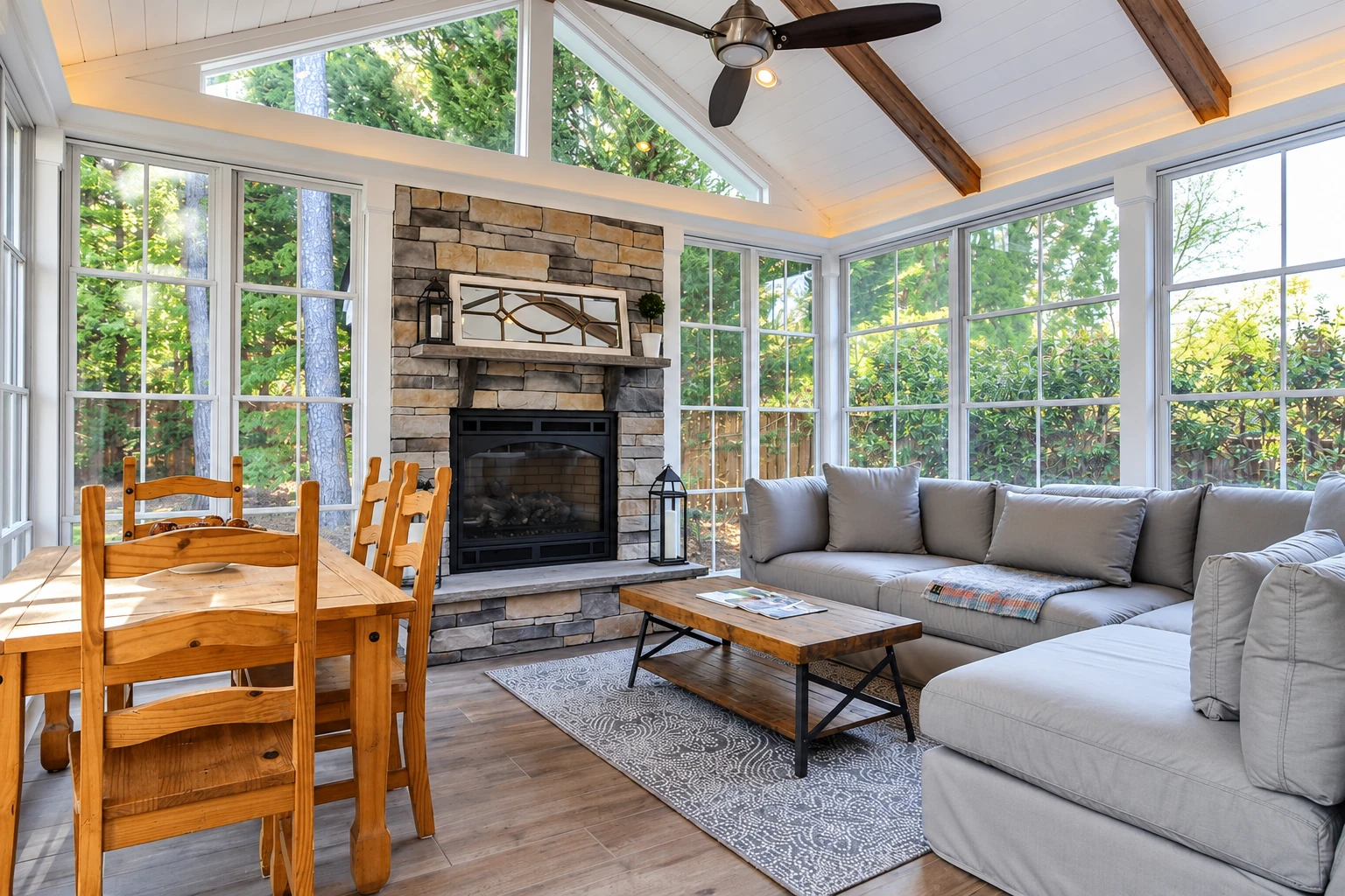 A bright three-season sunroom has large windows, a vaulted ceiling, a stone fireplace, and a dining table beside a gray sectional.